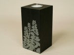 Czarny świecznik wykonany z drewna w kształcie prostopadłościanu. Na czołowej powierzchni motyw roślinny wykonany techniką sitodruku.